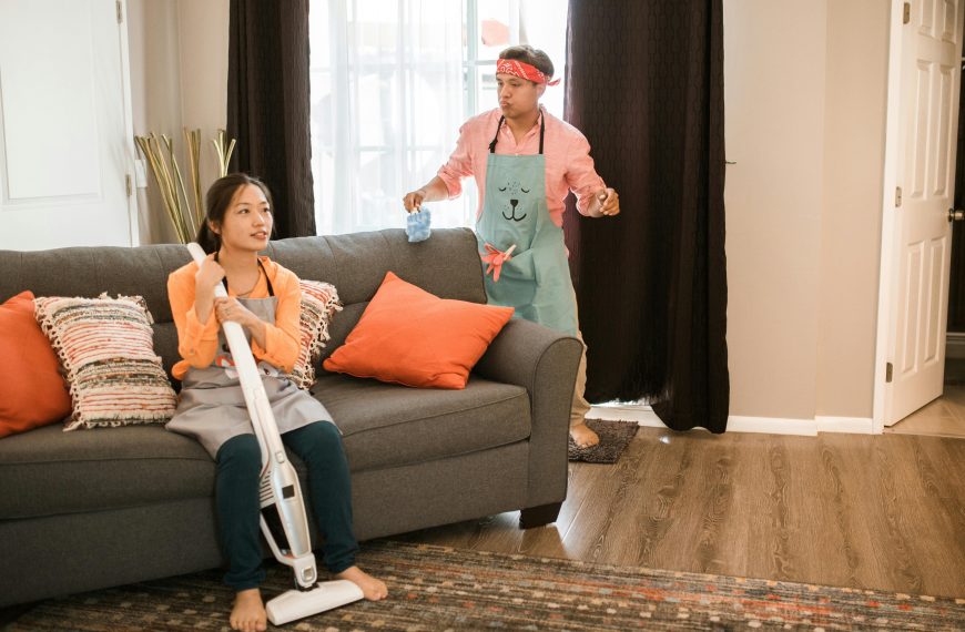 woman seated on a couch holding a vacuum and a man holding a duster